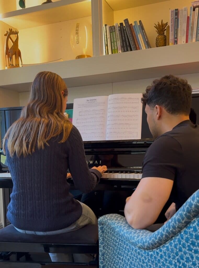 A piano teacher guiding an adult student at the keyboard.