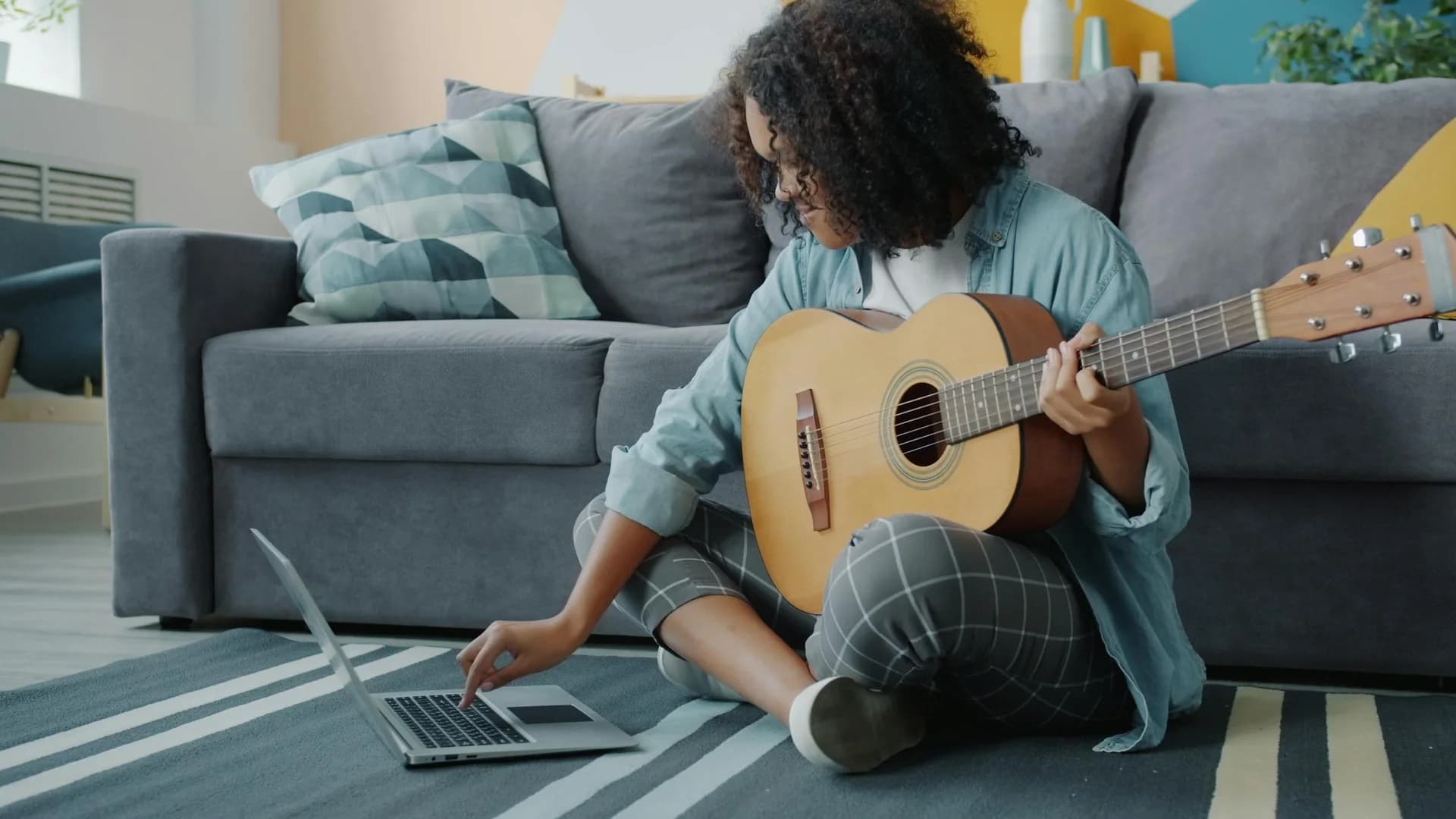 A student practising guitar at home with a laptop and notebook nearby.