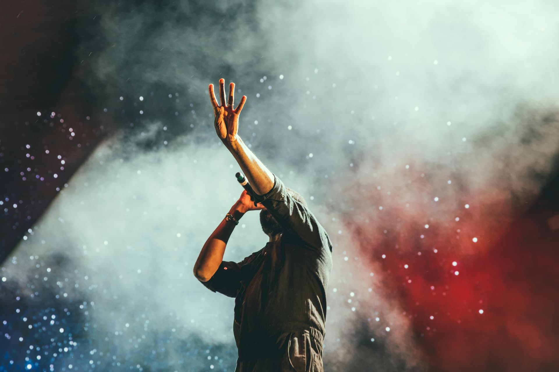 A vocalist on stage with an arm raised through smoke and stage light.