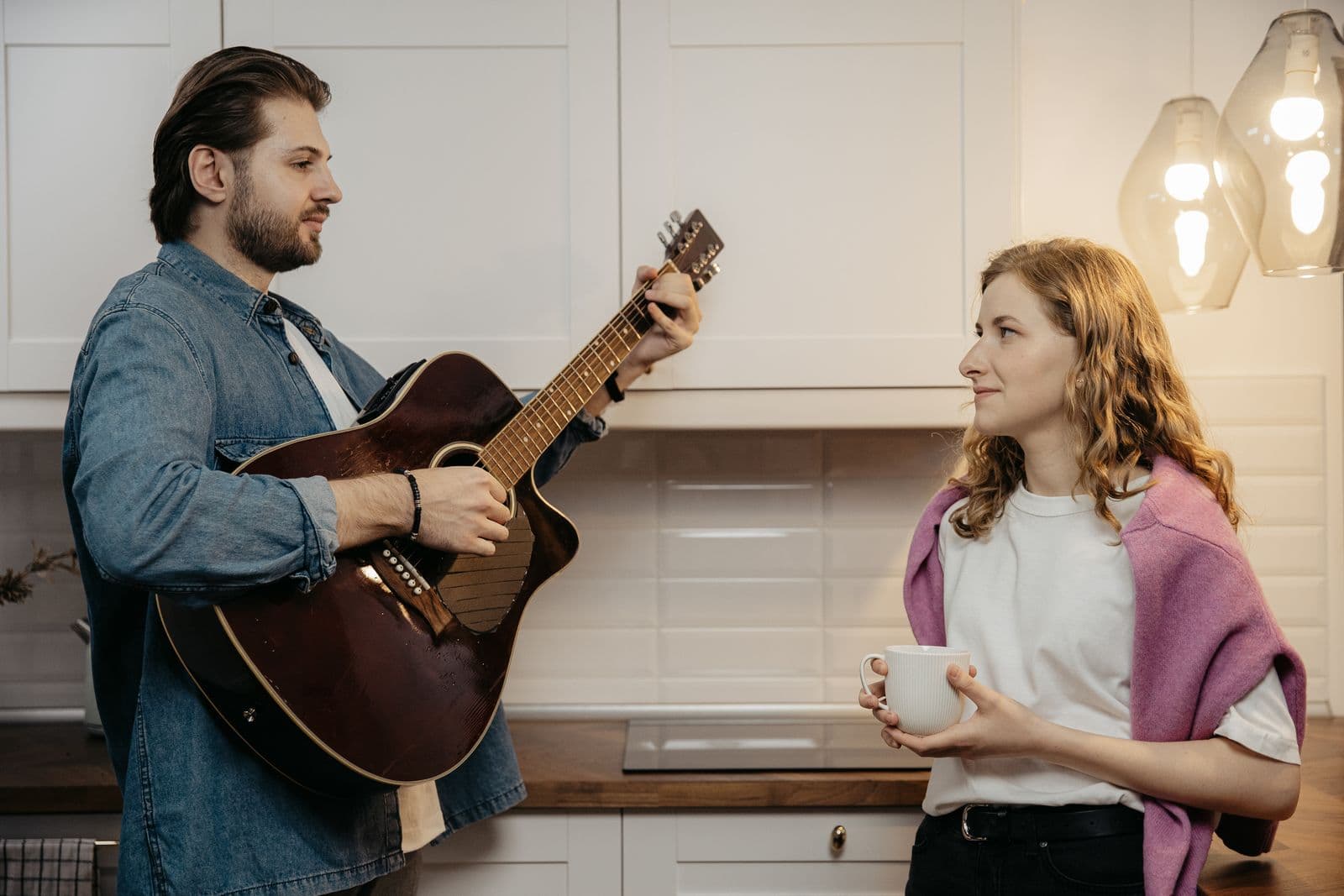 A guitarist playing at home while a friend listens over morning coffee.