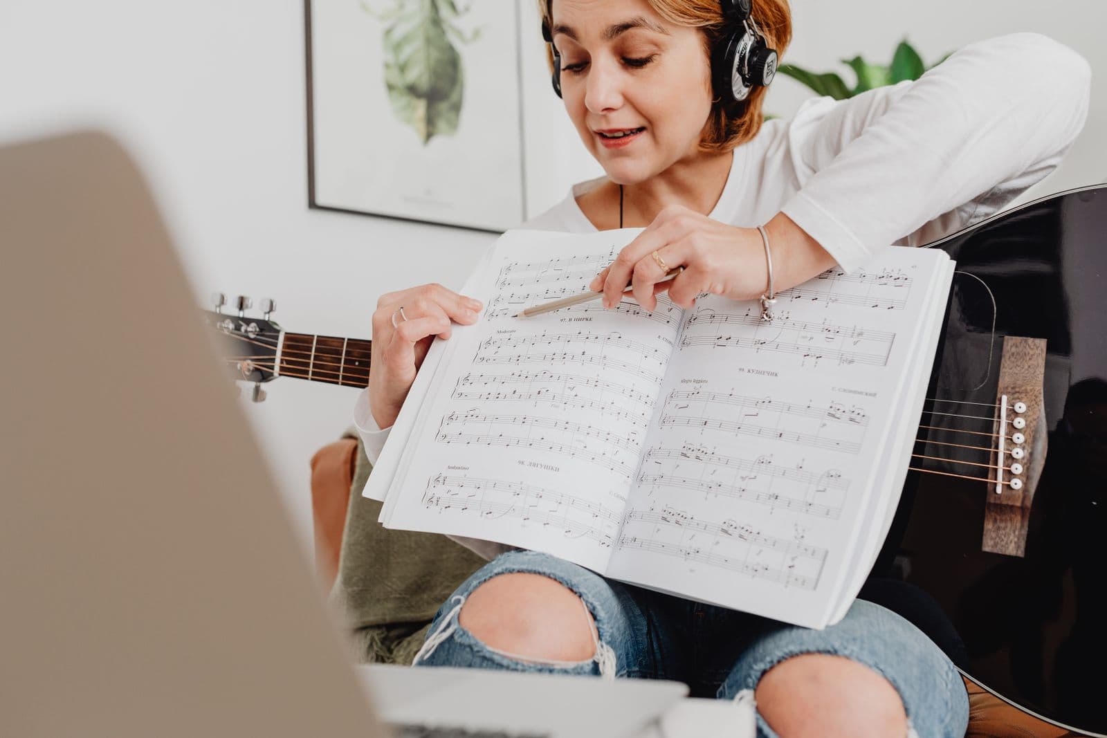 A teacher with headphones pointing out a passage in sheet music beside her laptop.