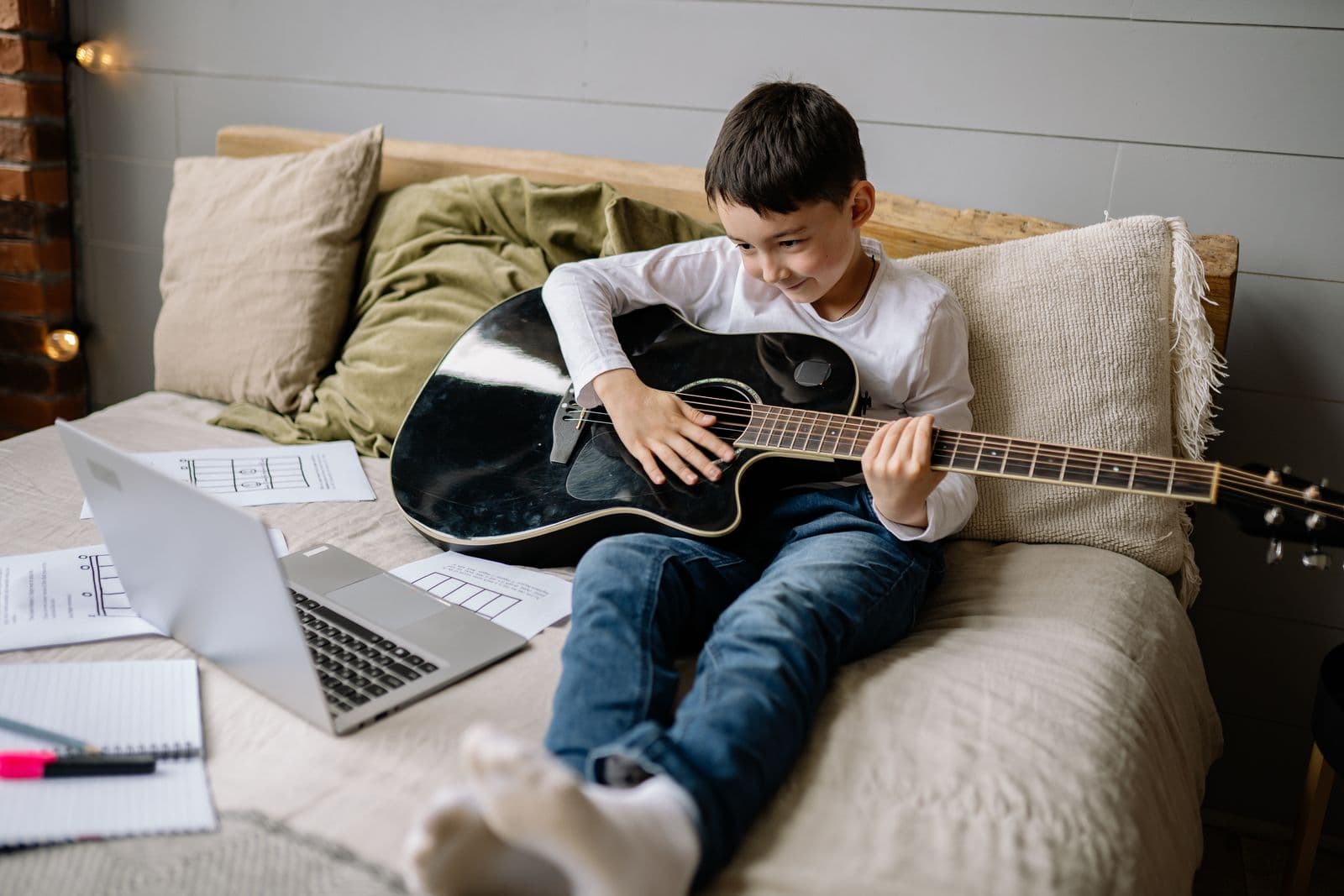 A young student with an acoustic guitar following an online lesson on his laptop.