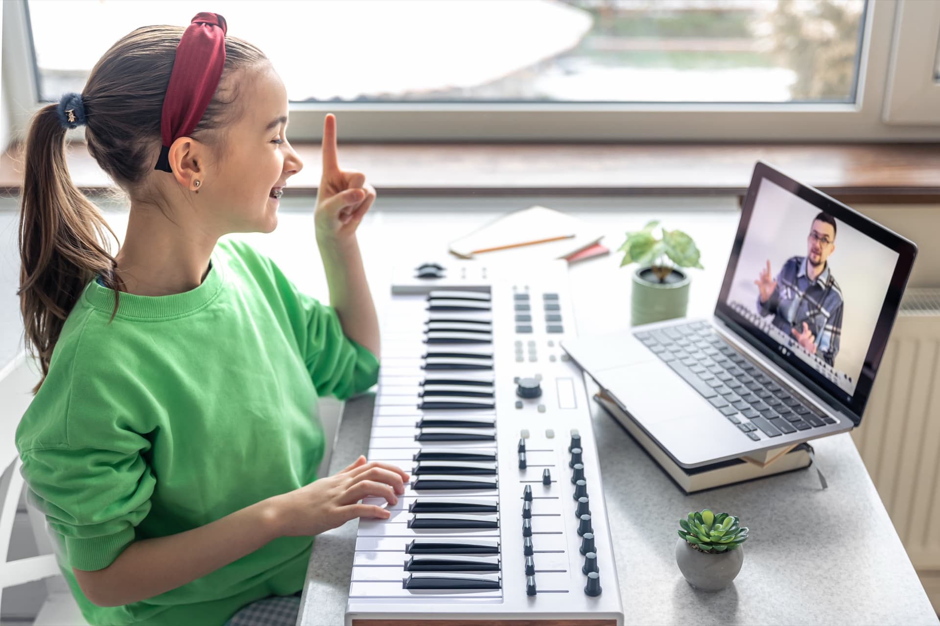 A young student at a keyboard taking an online lesson with her teacher on a laptop.