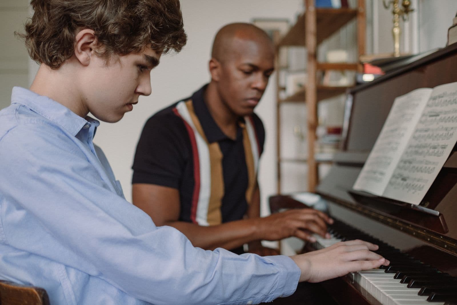 A piano teacher demonstrating at the keys while his student watches closely.