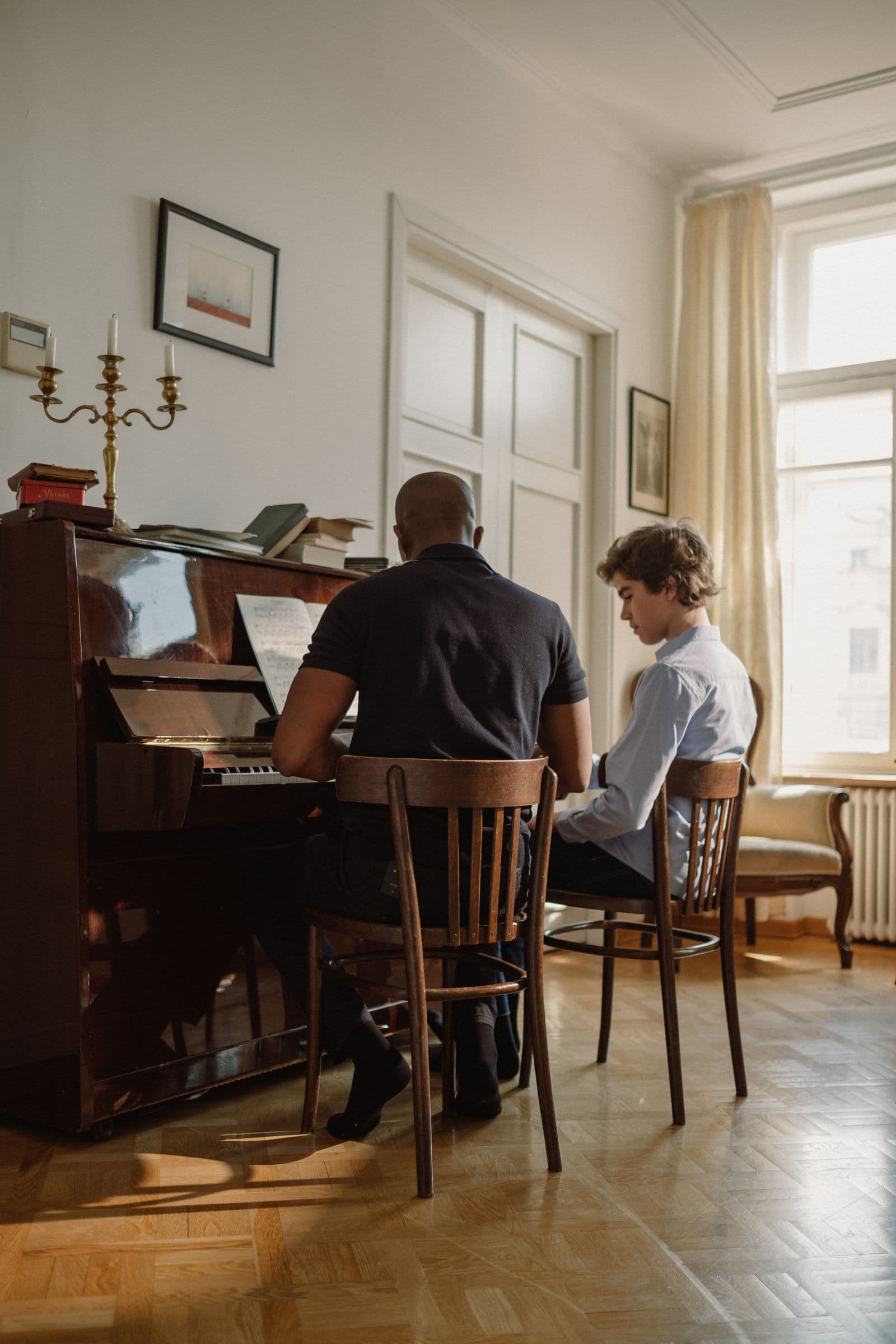 A piano teacher and student at an upright piano in golden window light.