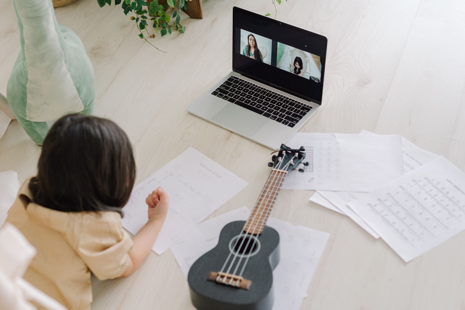 A young student taking an online music lesson on a laptop, with a ukulele and sheet music nearby.